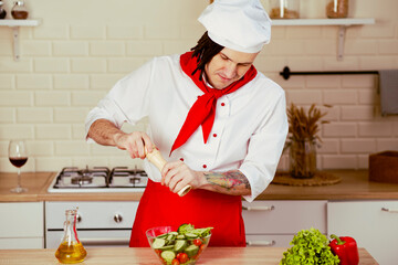 Young chef with dreadlocks peppering vegetable salad in kitchen. Smiling man in uniform preparing salad, grinding spices with mill.