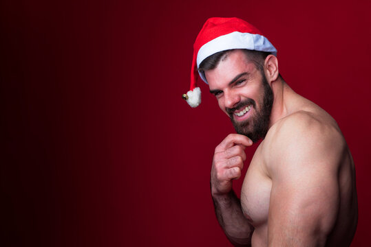 Sexy santa, young shirtless bodybuilder wearing a christmas hat, playing santa claus for holidays, red background studio portrait