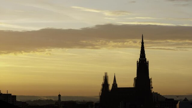 Time lapse shot with the silhouette of the Ulm minster in front of a yellow sky at morning while dawn and sunrise with some clouds