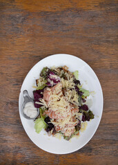 Top view of a Caesar salad with lettuce, parmesan cheese, bread croutons, salmon, bacon and Caesar dressing, in a white plate on the wooden table.