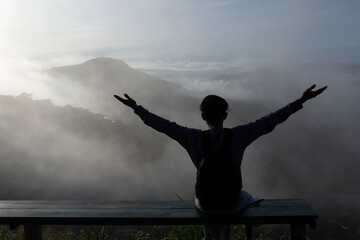 Fototapeta na wymiar Silhouette of young man sitting with happy and raise hands in the morning and mist cover mountain background
