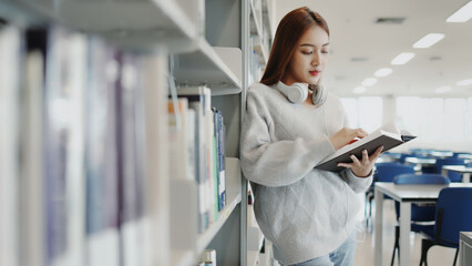 Asian young student girl in sweater searching the book from bookshelf in university or college...