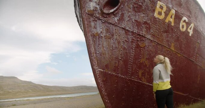 Young Woman Looking And Touching The Rusty Hull Of Gardar BA 64 Shipwreck On The Patreksfjordur Shoreline In Iceland. - wide