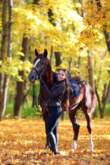 Equestrian model girl  walks with horse in the autumn woods