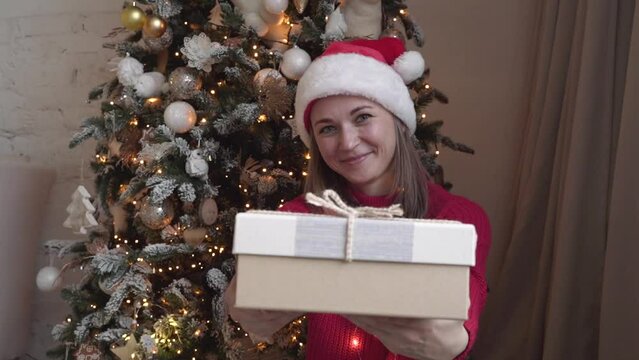 Woman wearing red sweater and Santa's cap presenting a gift near the tree