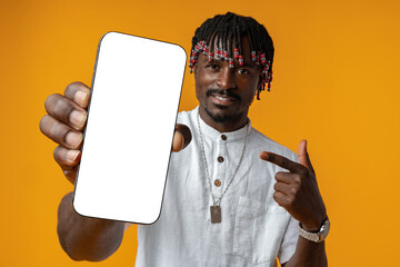 Excited young african man holding smartphone with white blank screen in hand on yellow studio...