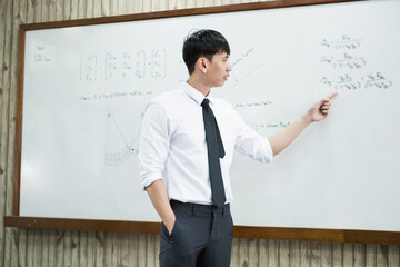 An Asian male teacher in a classroom near the whiteboard is writing and teaching a lesson with a...