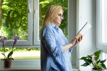 Pleasant blonde woman writer in glasses, stands near the window holding a paper notebook and reads.
