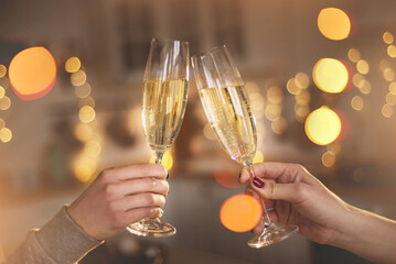 Two women clink glasses with champagne closeup  hands against the backdrop of a Christmas tree and lights. Lesbian couple celebrating. Women's hands hold thin crystal glasses against in the kitchen