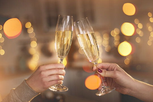 Two women clink glasses with champagne closeup  hands against the backdrop of a Christmas tree and lights. Lesbian couple celebrating. Women's hands hold thin crystal glasses against in the kitchen