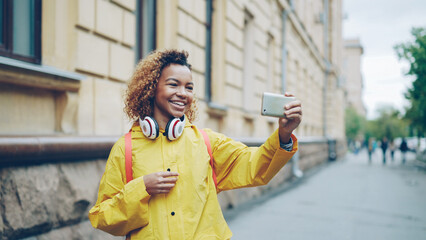 Fototapeta premium Cheerful African American girl is making video call, speaking and laughing holding smartphone and looking at screen. Modern technology, joyful communication and young people concept.