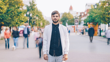 portrait of serious young man looking at camera standing in center of busy pedestrian street in summer wearing stylish clothing while many people are passing by. - 544810763