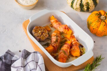 Baked pumpkin wrapped in bacon in a white ceramic baking dish on a light concrete background. Pumpkin recipes.