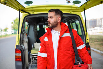 Fototapeta na wymiar Young man , a paramedic, standing at the rear of an ambulance, by the open doors. He is looking at the camera with a confident expression, smiling, carrying a medical trauma bag on his shoulder.