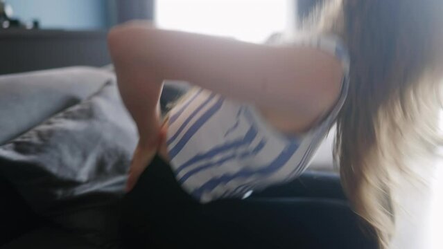 Caucasian woman sitting down on a couch at home experiences sudden back pain. Young adult female suffers from pain in her back descending on sofa. Girl rubbing back suffering from cramps in lumbar