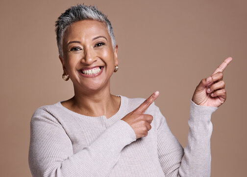 Woman, Smile And Happiness In Studio For Advertising Free Space, Promo Or Mockup On A Brown Background For Cosmetics, Makeup Or Fashion. Face Portrait Of A Mature Model Pointing Finger At Mock Up