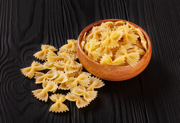 Uncooked farfalle rigate pasta in wooden bowl on black wooden background