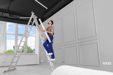 Electrician in uniform with ceiling lamp on metal ladder indoors. Space for text © New Africa