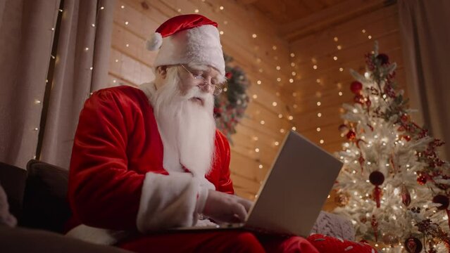 The real Santa Claus bottom view sits with a laptop on a couch with glasses and works at the laptop. Christmas and Christmas Eve Sonta Klaus works at home with garlands and a laptop in a decorated