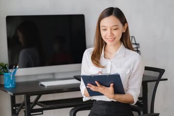 Attractive Asian businesswoman with a smile sits with a clipboard in her office, looking at the camera.