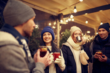 Happy smiling friends with cups of mulled wine having fun, spending time together at winter fair at evening time. Winter holidays, Christmas concept