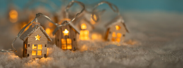 Fototapeta na wymiar Abstract Christmas Winter Panorama with Wooden Houses Christmas String Lights in Cold Snow Landscape and Glowing Golden Lights in Background. Panorama, Banner. Christmas or Energy themes.