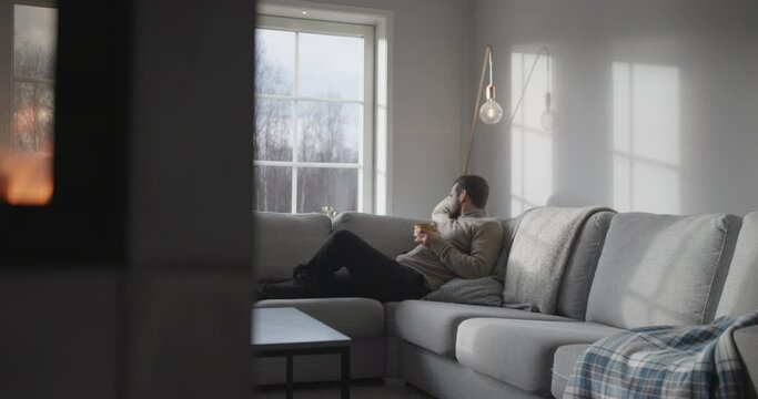 Man sits at home relaxing with cup of hot drink in sunlit room, fire burns in foreground, wide shot