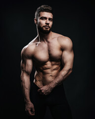 Slim muscular male model at black background. Fitness shirtless guy in black sport pants posing in studio. Man with six pack abs looking at camera. - obrazy, fototapety, plakaty