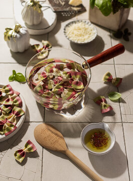 Pro cess of cooking colored beetroot and spinach farfalle are cooked in a transparent saucepan with basil, grated cheese on white tile background.Italian dish pasta on a sunny kitchen.