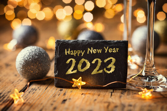 Happy New Year 2023 - Greeting Card - Black slate board with golden English text. Christmas decoration on rustic wood with Champagne glasses and golden  lights from fireworks in background