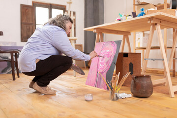 Mature artist woman paints on a canvas while squatting at her workshop as she develops her hobby