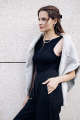 Young woman in black dress and sweater draped over the shoulders stands on street.