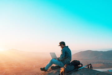 latino digital nomad sitting on top of the hill on a rock with his laptop working at sunset over...