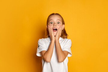 Portrait of little beautiful girl, child in white T-shirt posing in shock and excitement isolated over yellow studio background