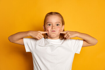 Portrait of little beautiful girl, child in white T-shirt posing, making funny faces isolated over yellow studio background. Grimacing