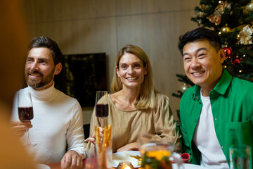 Happy young interracial people sitting at festive New Year's table. They are holding glasses of wine, talking, smiling, celebrating Christmas with friends.