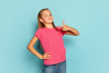 Portrait of little beautiful girl, child in pink T-shirt posing isolated over blue studio background. Proudly pointing at herself