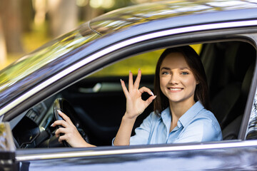 Attractive girl is sitting in a modern car. She is looking through window and showing okay sign.