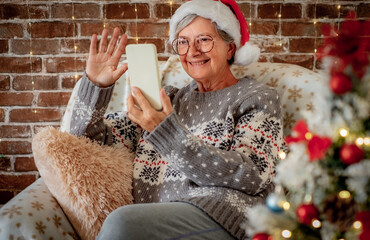 Portrait of beautiful senior woman in santa hat video chatting by mobile phone at Christmas time - elderly smiling lady sitting alone at home using wireless technology