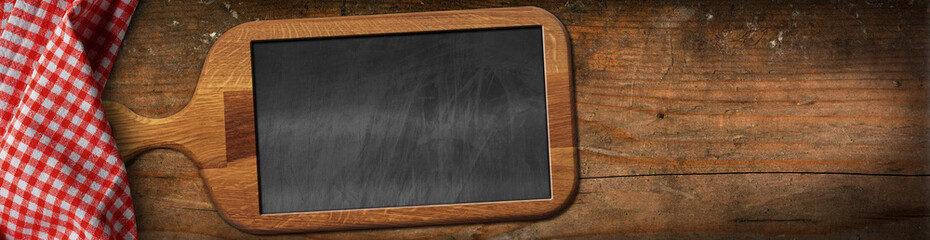 Old wooden cutting board with a blank chalkboard inside, on a wooden table with a red and white checkered tablecloth. Template for recipes or food and drink menu.