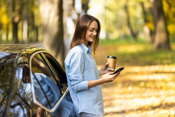 Young woman with a mobile phone and coffee cup standing near charging electric car. Vehicle...