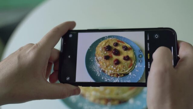 Woman makes photo of cooked pancakes with raspberries and sugar powder served on blue plate. Female hands hold smartphone with food on screen closeup