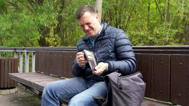 Millennial man takes out of bag and checks an empty wallet without money. Mature male has no money in wallet, stressed and depressed on bench at park. Concept of poverty. 