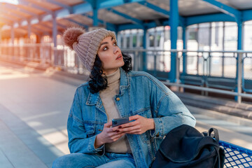 a young woman in a denim jacket is talking on the phone and waiting for a tram at the stop...