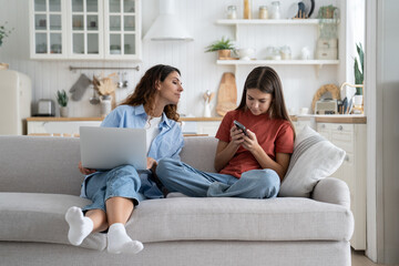 Teen girl child daughter and young mother using gadgets together, sitting on sofa at home. Curious...