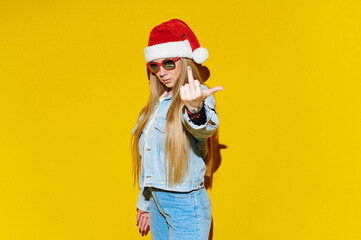 Young Christmas woman in Santa Claus hat and sunglasses showing middle finger over yellow background. Feeling provocative, aggressive and obscene