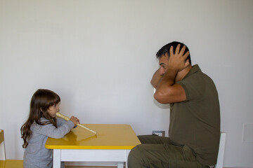 Little girl playing the flute and dad plugging his ears. Music lessons of a little girl learning to...