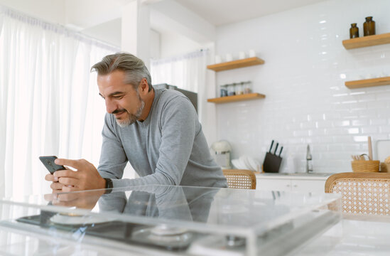 Smiling Middle age Caucasian man text messaging, chat social media through smartphone while sit in kitchen at home. Senior Male using apps ordering delivery food on mobile phone relax at kitchen room.