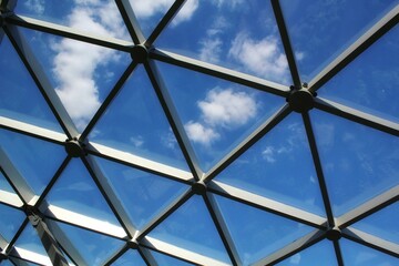 Transparent glass dome roof of modern design style pavilion building with geometric metal construction pattern. View of blue sky and clouds. Abstract structure triangle high-tech architecture window.