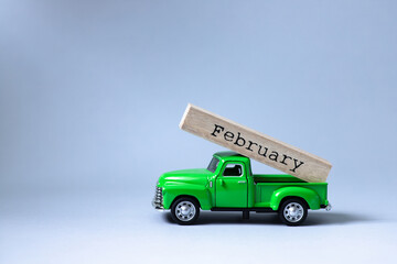 Green retro toy car carrying part of a wooden calendar with the month of February on grey background. Christmas and New Year celebration concept. Greeting card. Copy space, selective focus